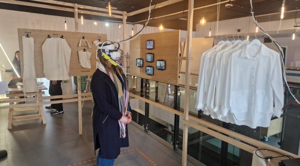Helena stands in front of rail of redesigned shirts while wearing a VR headset and exploring the future of fashion