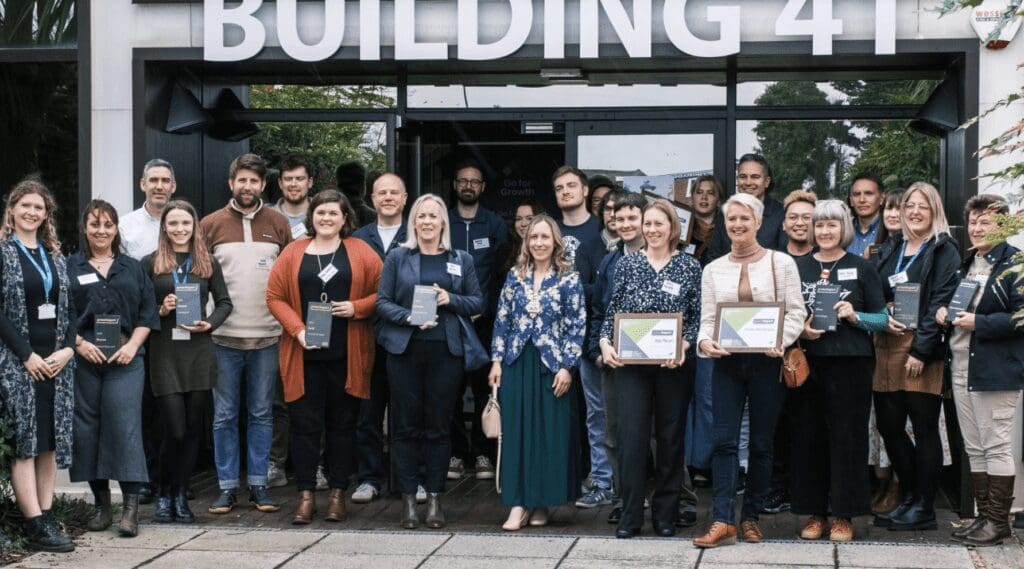 A group of people smile together outside a building as they hold awards certificates.