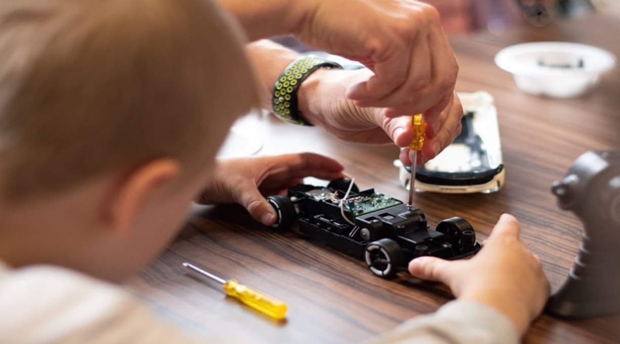 A child and an adult sit at a desk repairing a remote control car that has been disassembled.