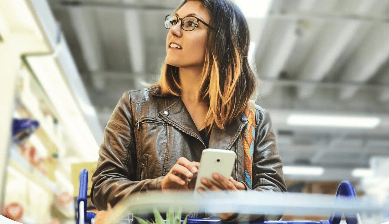 Woman looks up at supermarket shelf while holding a phone.