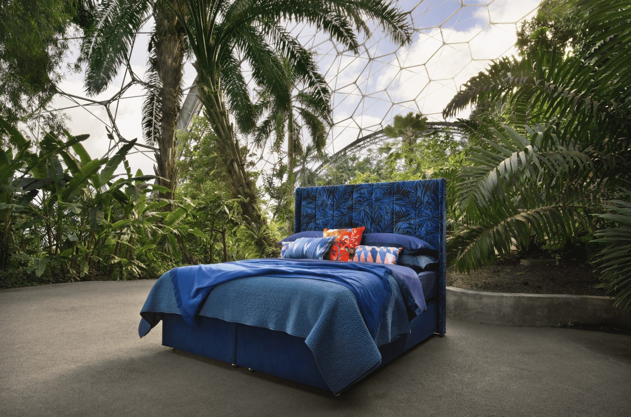 a blue bed with deep blue bedding is centred among tropical plants and trees found in the Eden Project biome