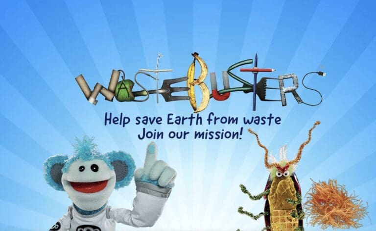 A blue monkey puppet stands on blue background and points upwards at Wastebuster sign made from arranged waste.