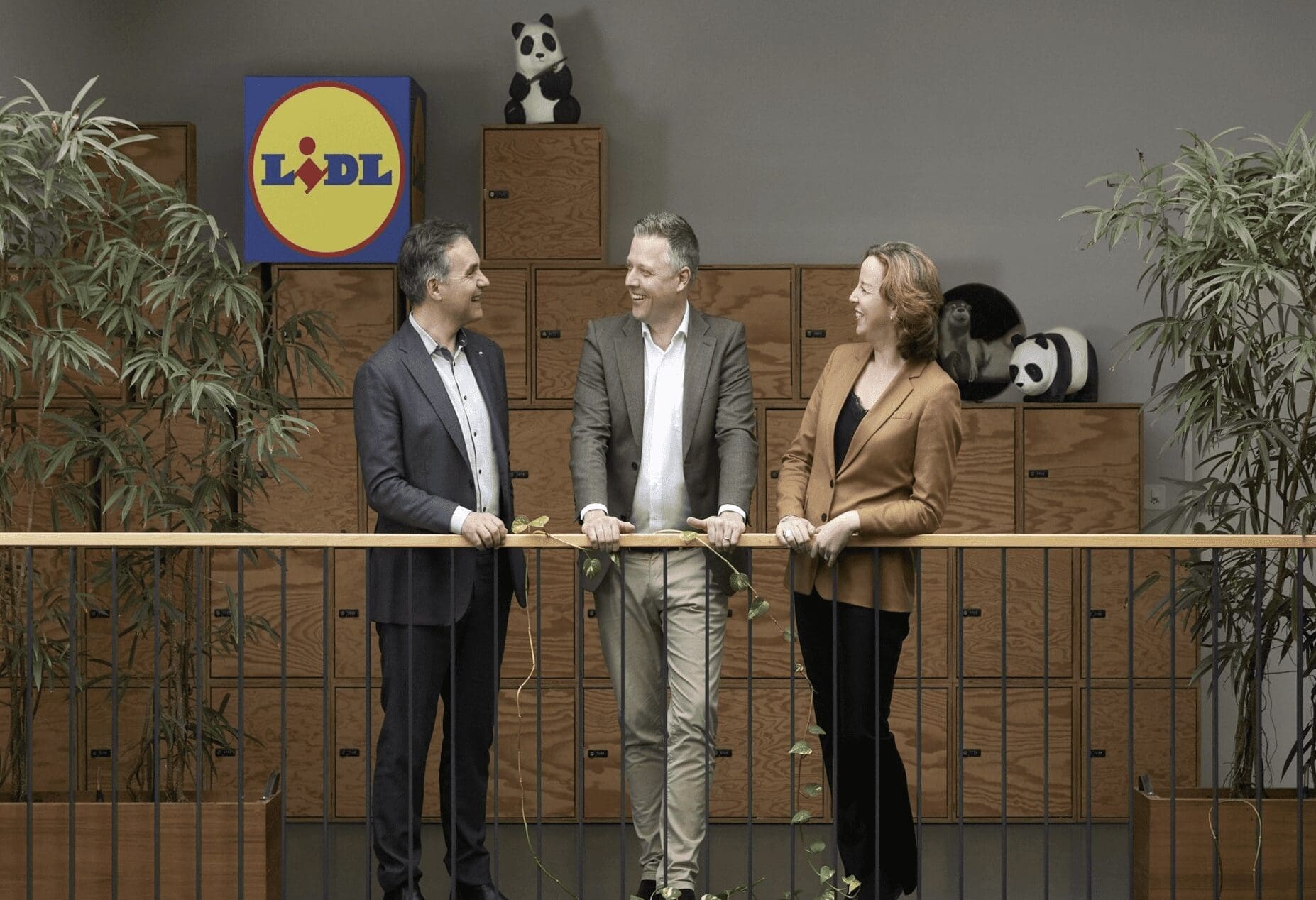 two men and a woman stand along a balcony in front of a stack of boxes with the Lidl logo and a cuddly toy panda on top.