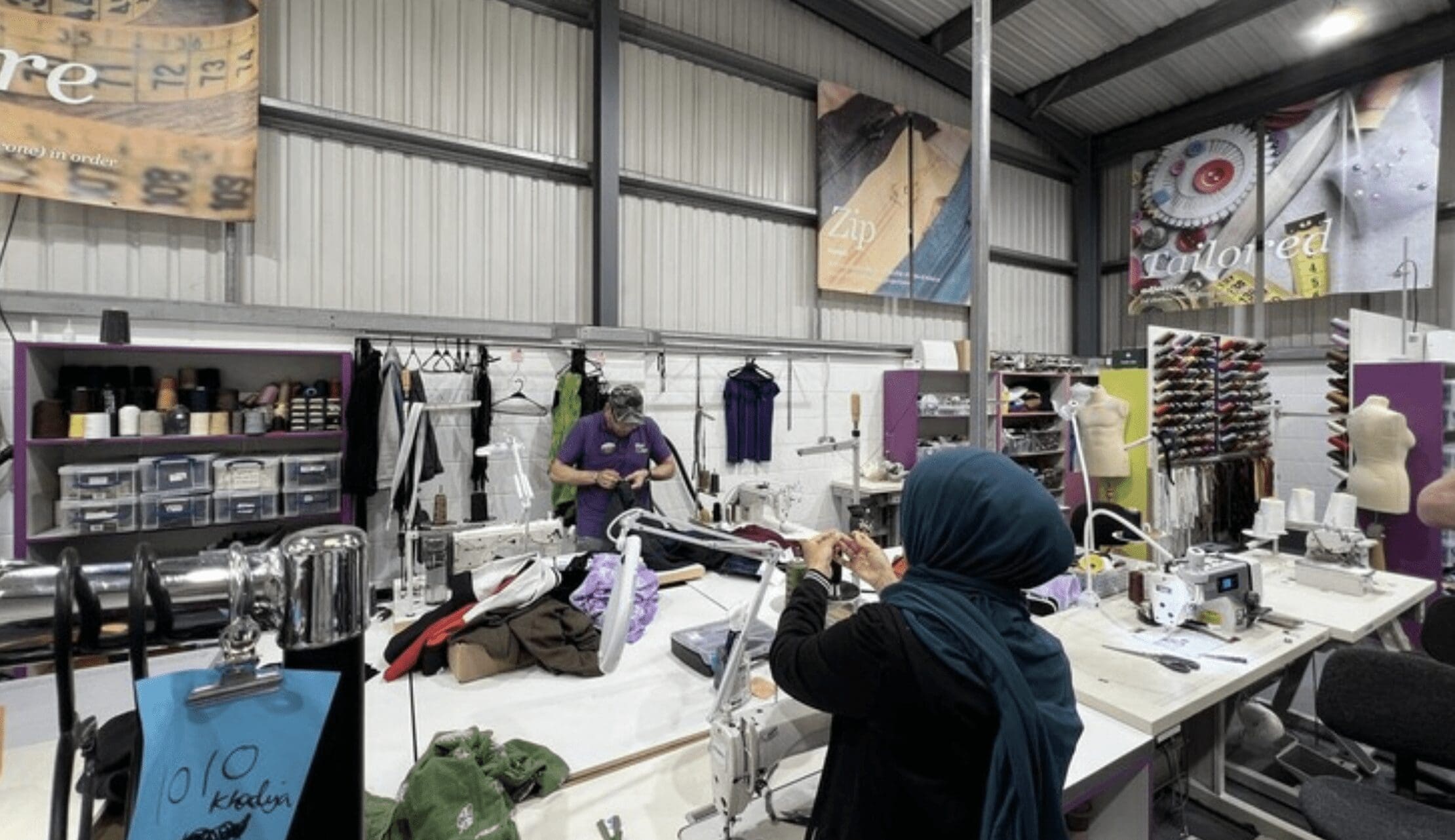 Two people at work in a clothing repair workshop with textiles and sewing machines around them.