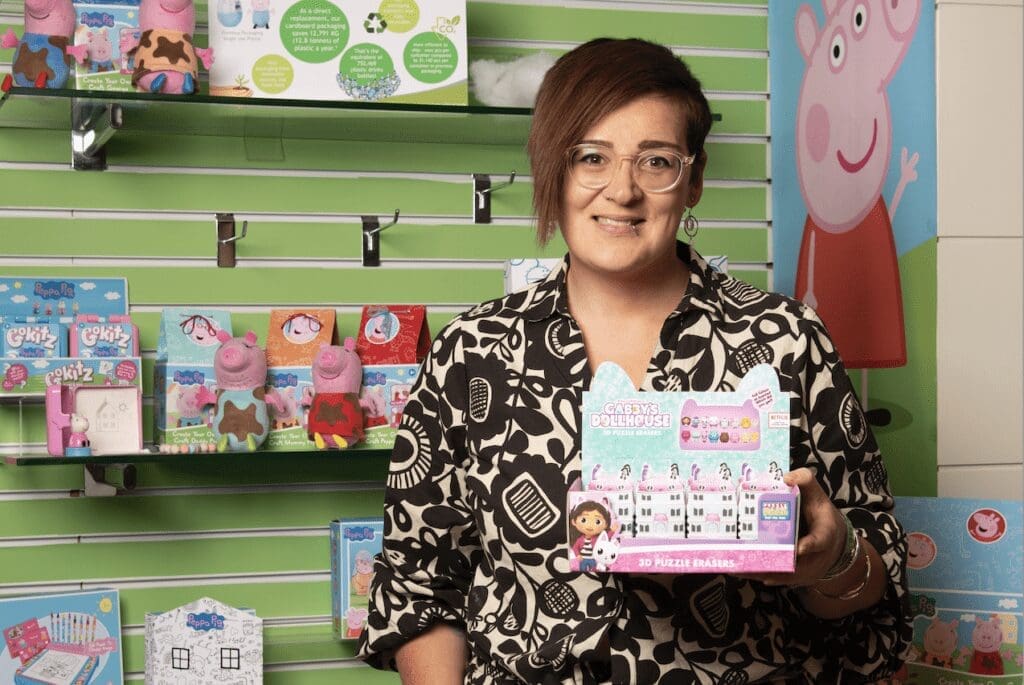 A woman stands in front of a green wall with a Peppa pig motif while she holds a box of toys