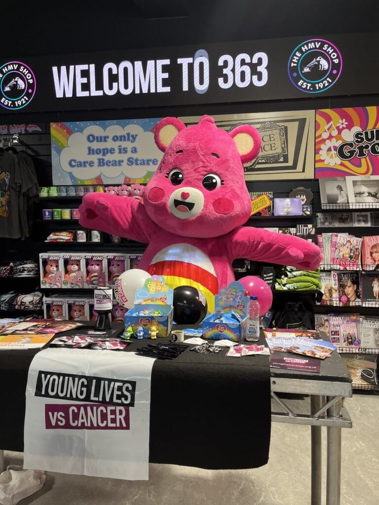 A Care Bear standing in HMV in front of a table with objects for Young Lives vs Cancer