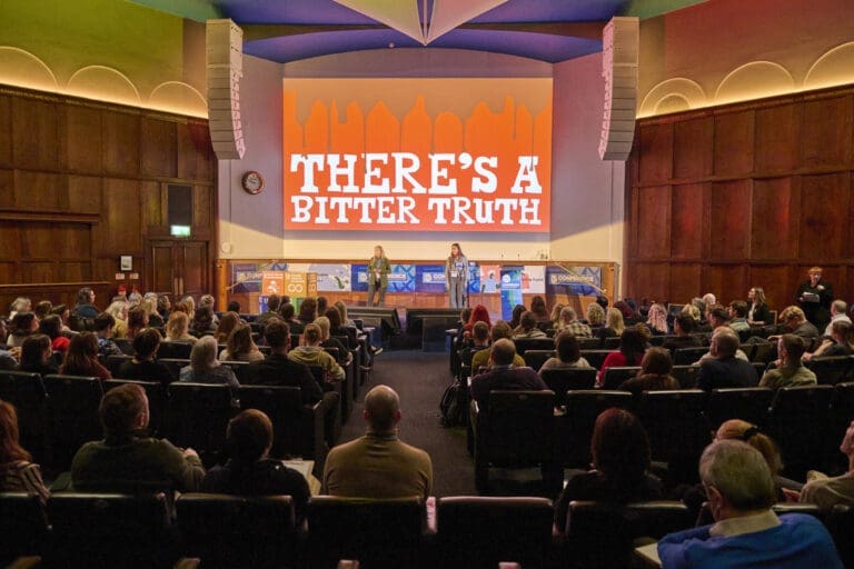 Two people stand on stage in front of an audience, with a screen behind them reading "There's a bitter truth"