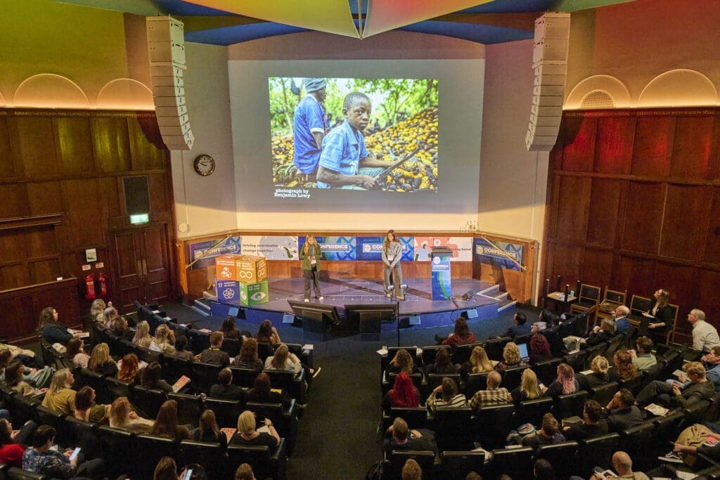 Two people on stage in front of an audience with a photo of a child on a cocoa farm on the screen behind
