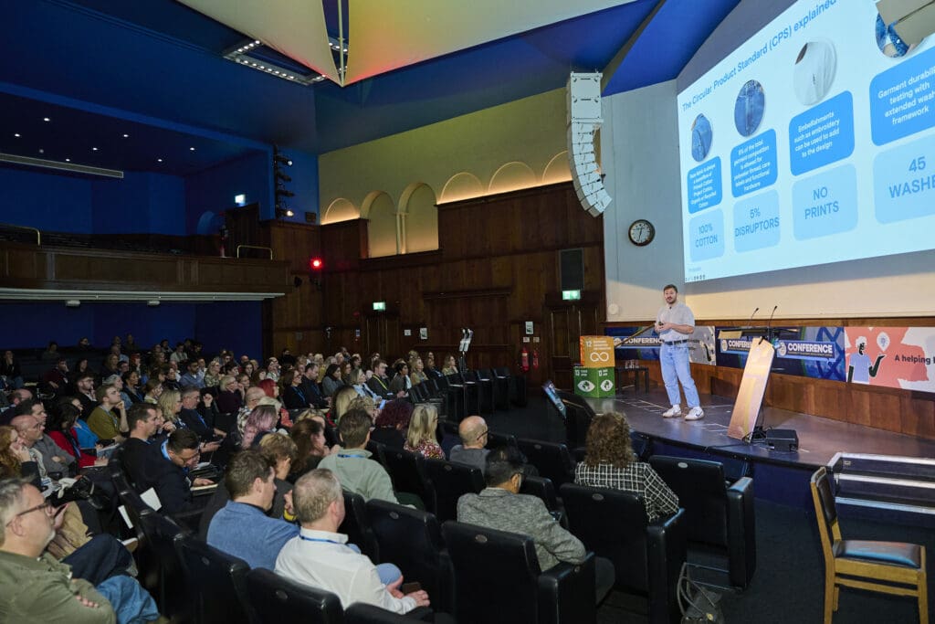 Man looks out over an audience on stage with a presentation on the screen behind him.
