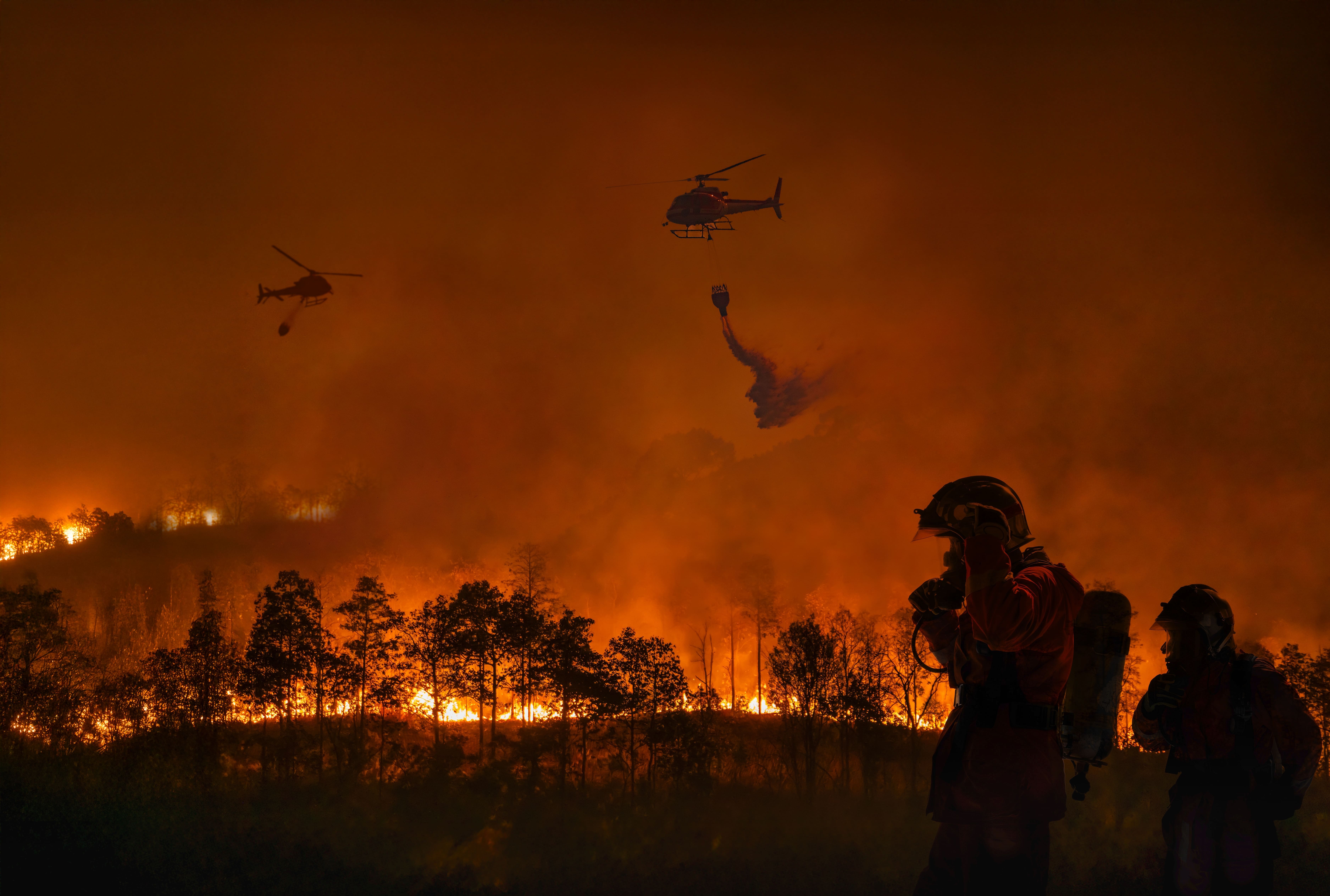 Nighttime shot of helicopters dropping water on a burning landscape, with firefighters silhouetted in the foreground.