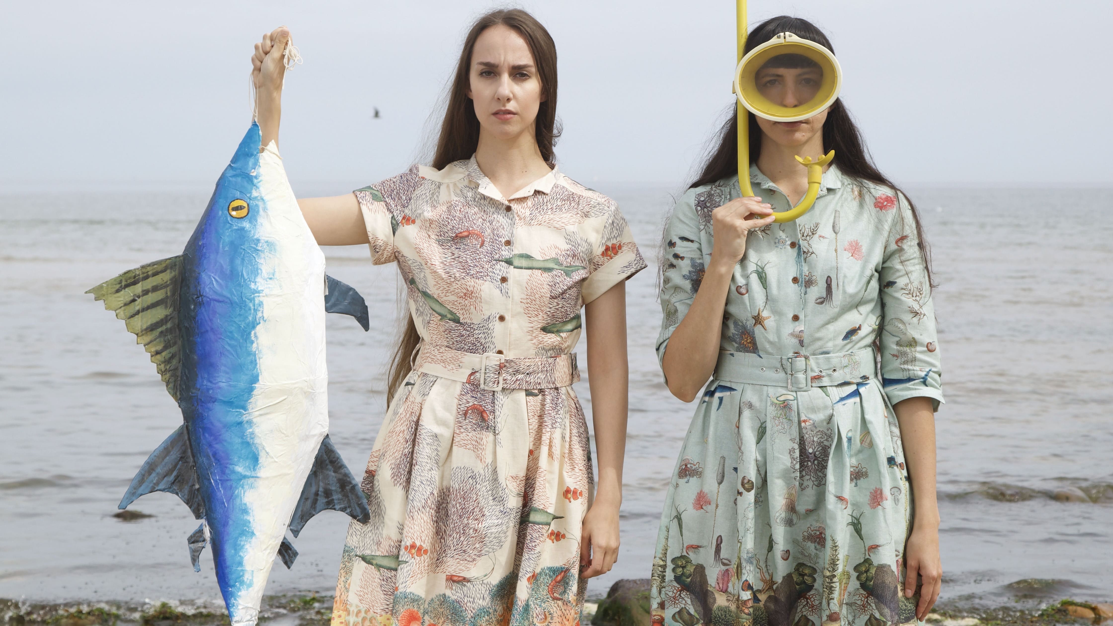 Two women stand in front of the sea shore in printed dresses, one wearing a snorkel and mask, the other holding up a fish